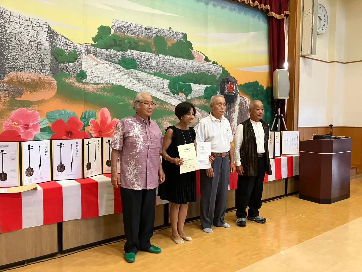 Quatre personnes posent devant une grande fresque colorée représentant un paysage d’Okinawa, lors d’une cérémonie de remise d’un sanshin destiné au Musée national de la Musique en France.