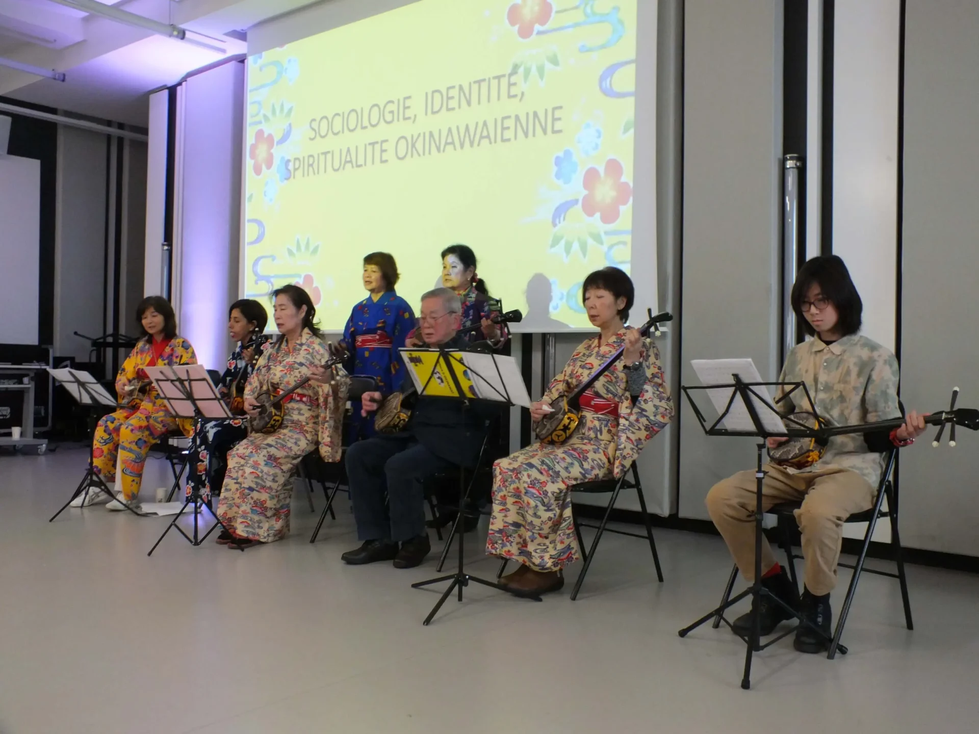 Groupe de musiciens en tenue traditionnelle okinawaïenne jouant du sanshin assis devant un écran de projection portant le titre « Sociologie, identité, spiritualité okinawaienne ».